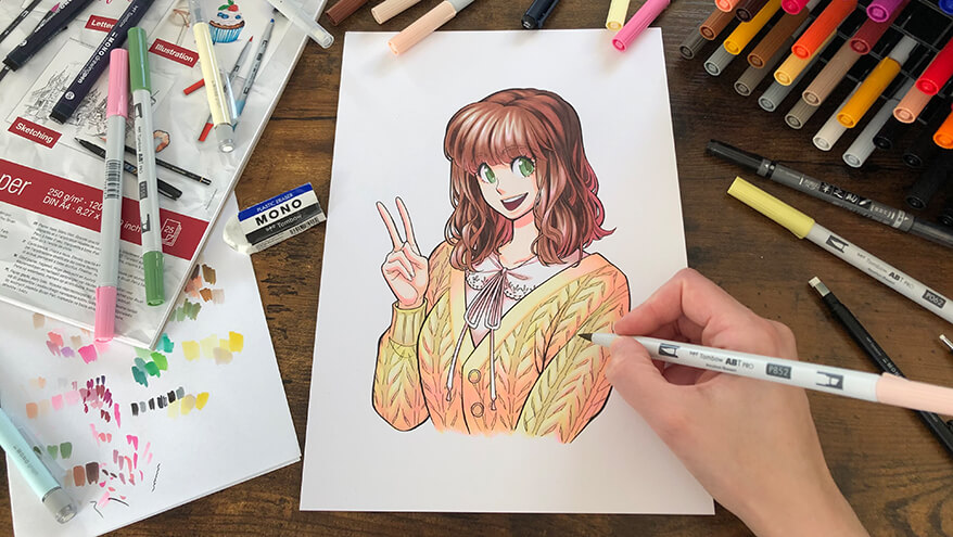 Jemand koloriert mit einem Marker ein Manga. Dieses zeigt eine junge Frau mit braunen Haaren und einem gelben Pulli, die lachend die rechte Hand mit dem Peace-Zeichen hochhält. Drumherum sind viele Zeichenmaterialien der Marke Tombow verteilt. Foto: Reyhan Yildirim