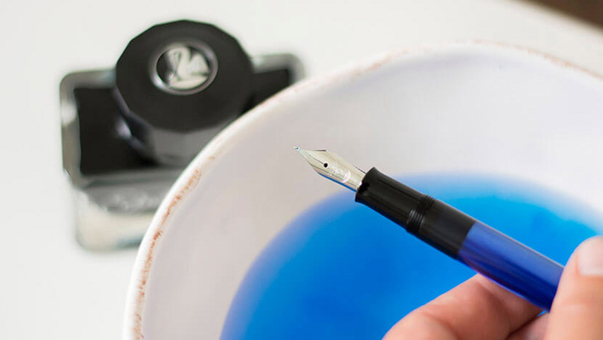 Ein Füller wird mit Wasser gereinigt. Das mit Tinte verfärbte Wasser ist zu sehen sowie ein Tintenfass der Marke Pelikan.