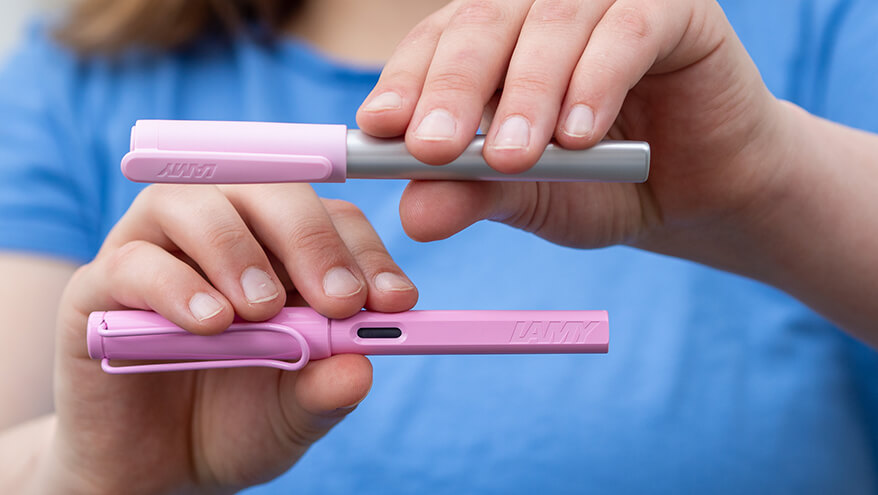 Ein Teenager-Mädchen im blauen T-Shirt hält zwei rosa Füller von Lamy zwischen ihren Fingern waagerecht in die Kamera.